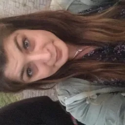 Uśmiechnięta kobieta z długimi brązowymi włosami, w bluzce w kwiaty i zielonej kurtce, pozuje do selfie na tle chodnika i fragmentu zieleni.