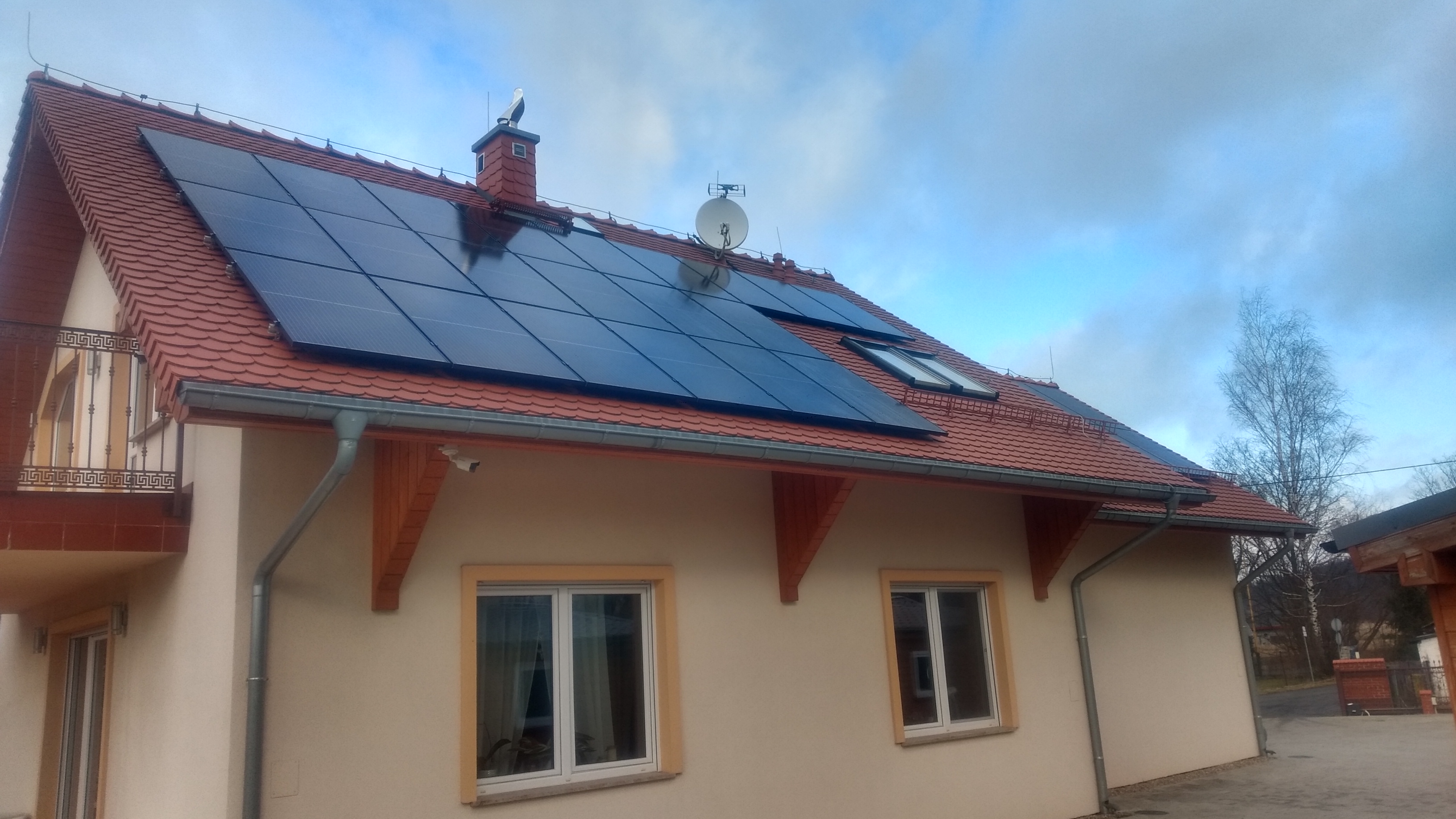 Dom jednorodzinny z panelami fotowoltaicznymi na dachu z czerwonej dachówki, widoczna antena satelitarna i ozdobny komin ze stojącym bocianem, pochmurne niebo.