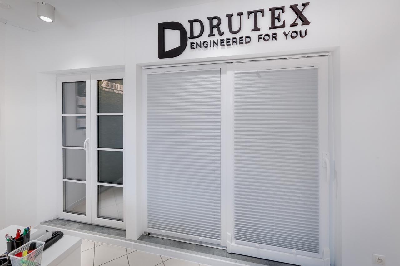 Białe okno PCV z roletą plisowaną oraz okno z podziałami, prezentowane w salonie firmowym z logo DRUTEX na ścianie.