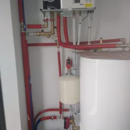 Instalacja kotła grzewczego z zasobnikiem ciepłej wody, widoczne rury w izolacji termicznej, zawory i podłączenia hydrauliczne na tle białej ściany.