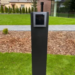 Nowoczesny wideofon na czarnym słupku wkomponowany w architekturę posesji z widocznym budynkiem z czerwonej cegły i starą zabudową w tle.