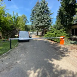 Szlaban automatyczny z pomarańczową obudową i białym ramieniem blokuje wjazd na teren posesji z zaparkowanym białym vanem w tle.