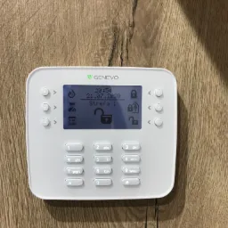 Biały panel sterowania alarmu Genevo z podświetlanym ekranem LCD, pokazujący datę 21.07.2020, godzinę 20:54 i status odblokowanej strefy, zamontowany na drewnianej ścianie.