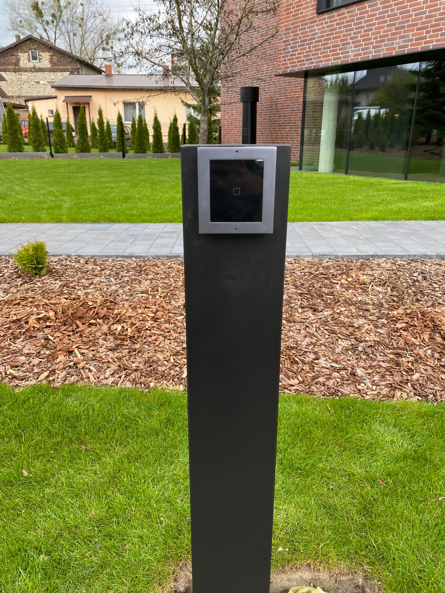 Nowoczesny wideofon na czarnym słupku wkomponowany w architekturę posesji z widocznym budynkiem z czerwonej cegły i starą zabudową w tle.