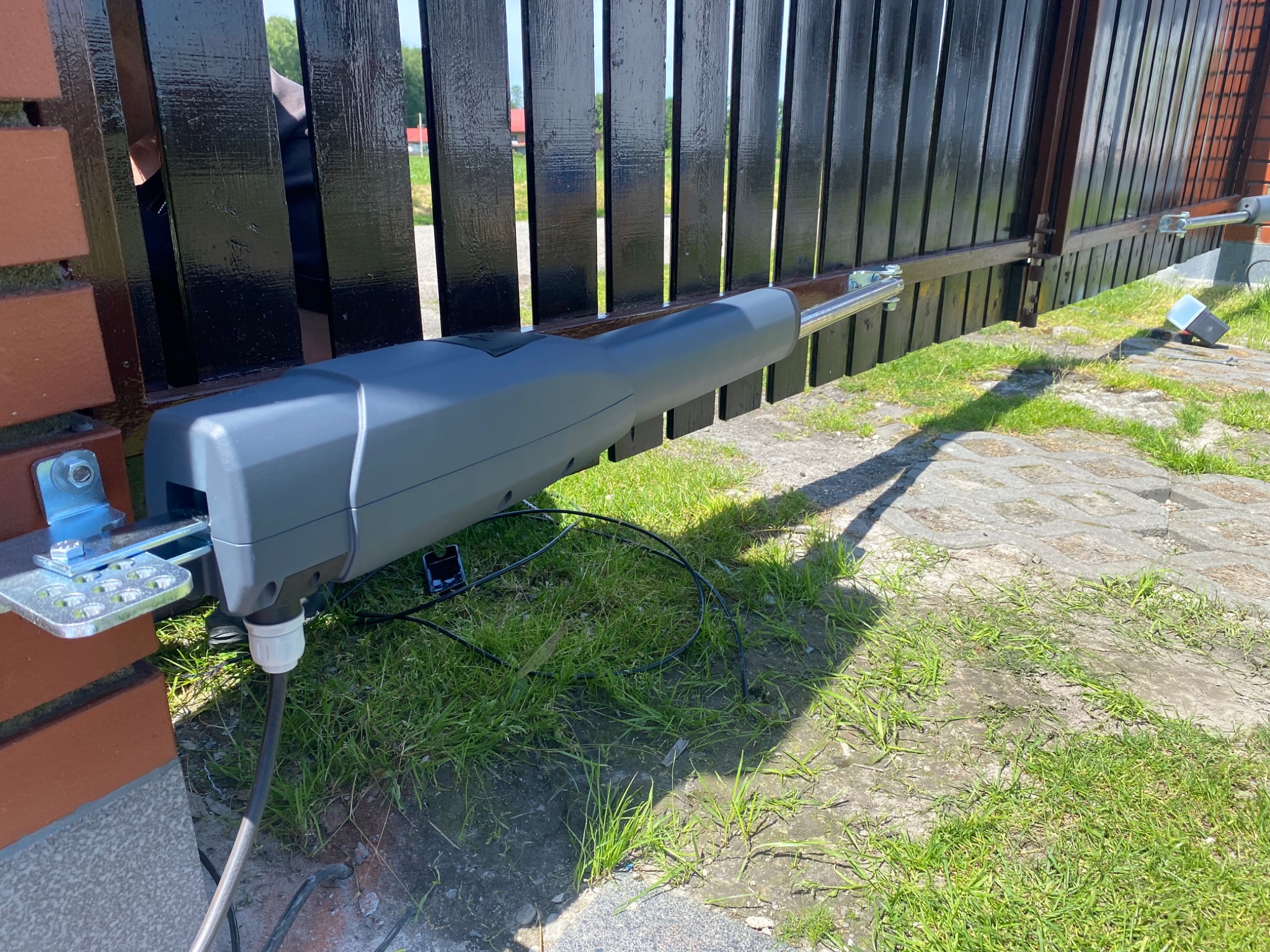 Szary siłownik bramy skrzydłowej zamontowany na czarnym ogrodzeniu z ceglanym słupkiem, widoczne przewody i telefon obok, trawa i kostka brukowa w tle.