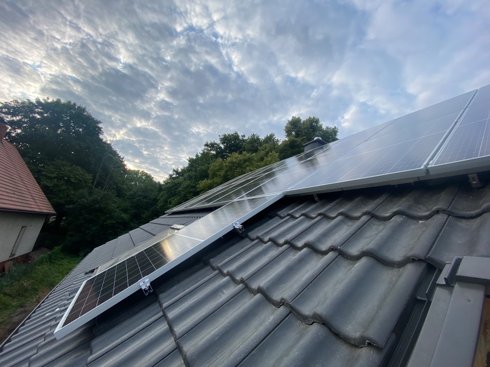 Panele fotowoltaiczne zamontowane na ciemnej dachówce, widok z dołu pod kątem, na tle pochmurnego nieba i drzew