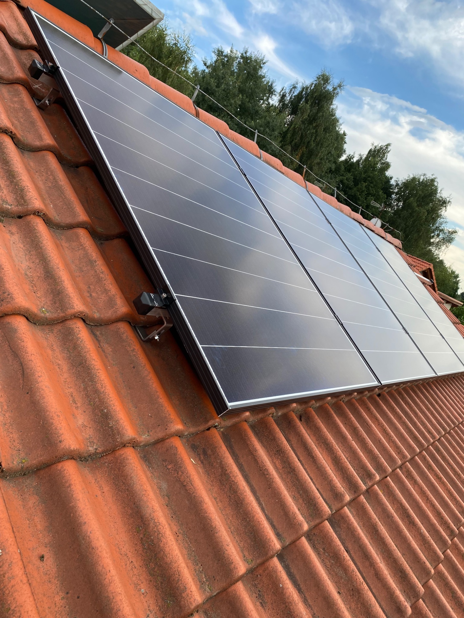 Panele fotowoltaiczne zamontowane na czerwonym, ceramicznym dachu z widocznymi mocowaniami, na tle błękitnego nieba z chmurami i zieleni drzew.