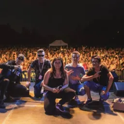 Pięcioosobowy zespół na scenie podczas koncertu plenerowego, w tle rozentuzjazmowany tłum fanów z podniesionymi rękami.