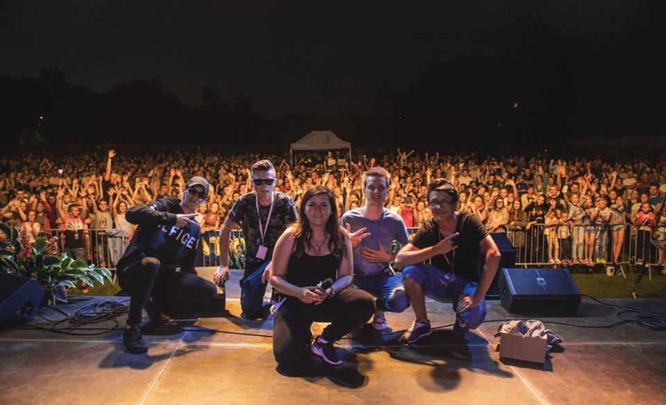 Pięcioosobowy zespół na scenie podczas koncertu plenerowego, w tle rozentuzjazmowany tłum fanów z podniesionymi rękami.