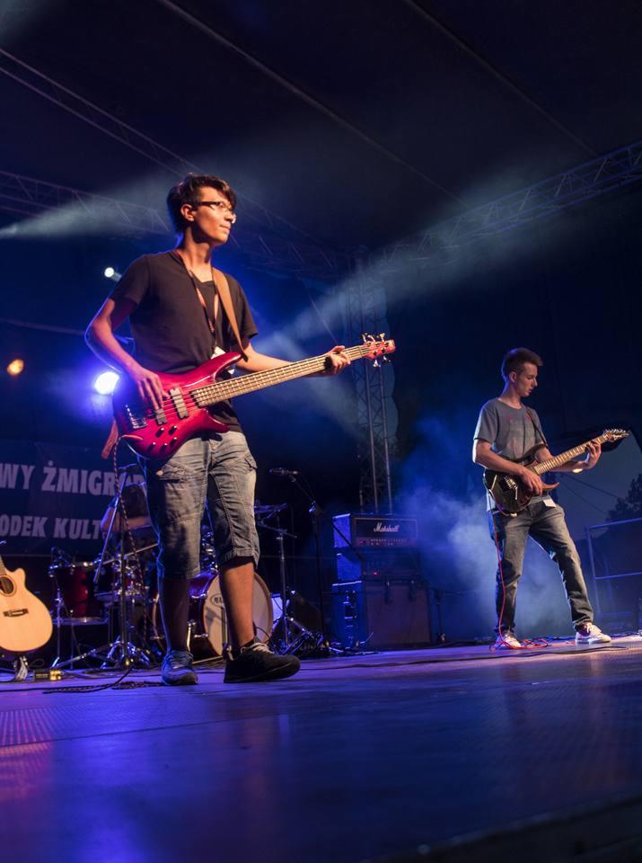 Dwóch młodych muzyków gra na scenie podczas koncertu plenerowego, w tle perkusja i wzmacniacze Marshall, niebieskie oświetlenie sceniczne.