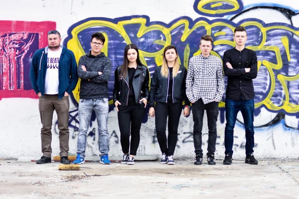 Sześcioosobowa grupa młodych ludzi pozuje na tle ściany pokrytej kolorowymi graffiti w stylu miejskim.