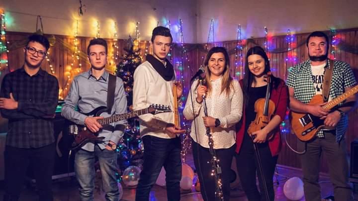 Sześcioosobowy zespół muzyczny z gitarami i skrzypcami, występujący na tle światełek i choinki, jeden z muzyków ma koszulkę z napisem BEAT.