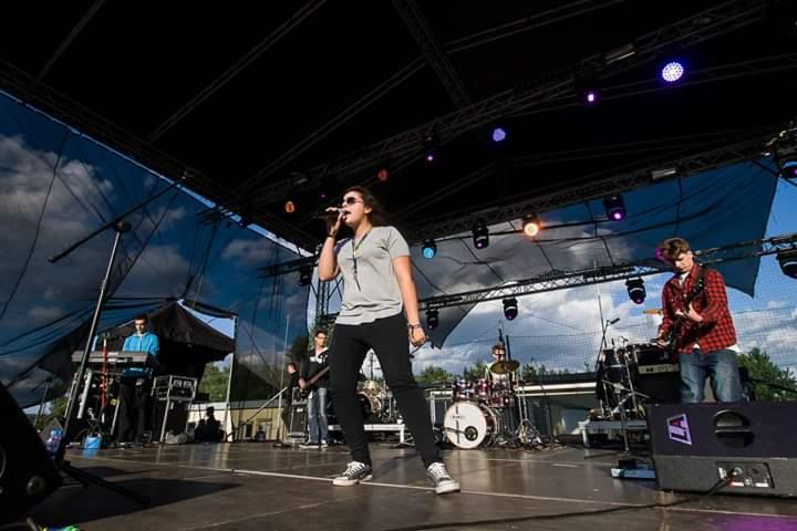 Wokalistka śpiewa do mikrofonu na scenie podczas występu plenerowego, w tle zespół z keyboardem, perkusją i gitarą.