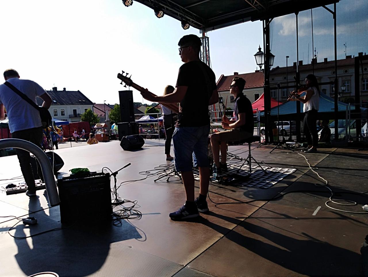 Występ na scenie plenerowej: basista, perkusista i skrzypaczka podczas próby dźwiękowej, w tle budynki miejskie i stragany.