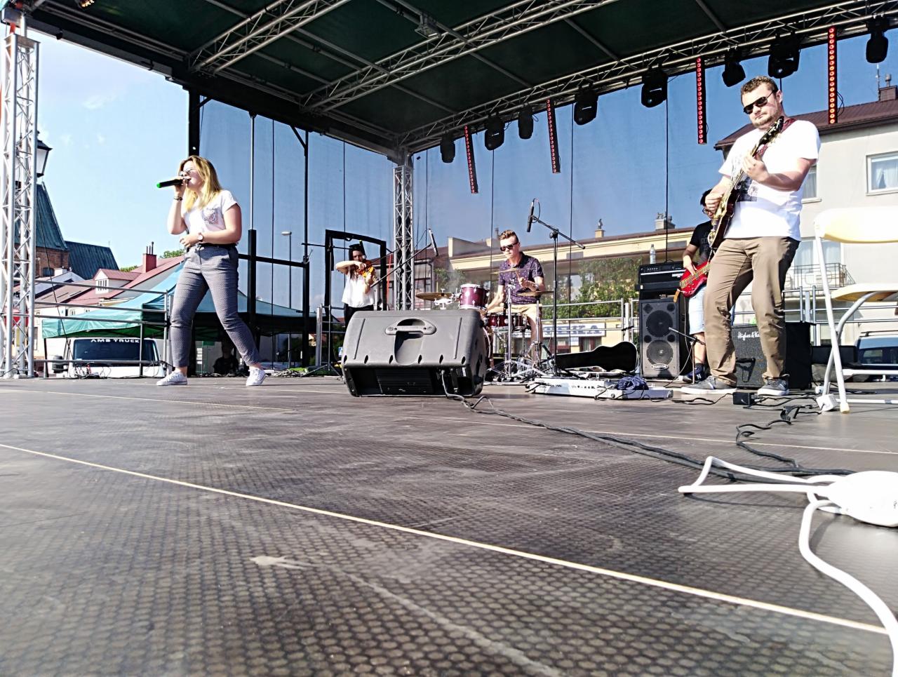 Występujący na scenie muzycy: wokalistka z mikrofonem, gitarzysta w okularach słonecznych, skrzypek i perkusista podczas koncertu plenerowego w słoneczny dzień.