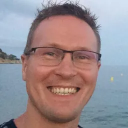 Portret uśmiechniętego mężczyzny w okularach na tle morza i skalistego brzegu w pochmurny dzień. Widoczne białe zęby i krótko ostrzyżone włosy.