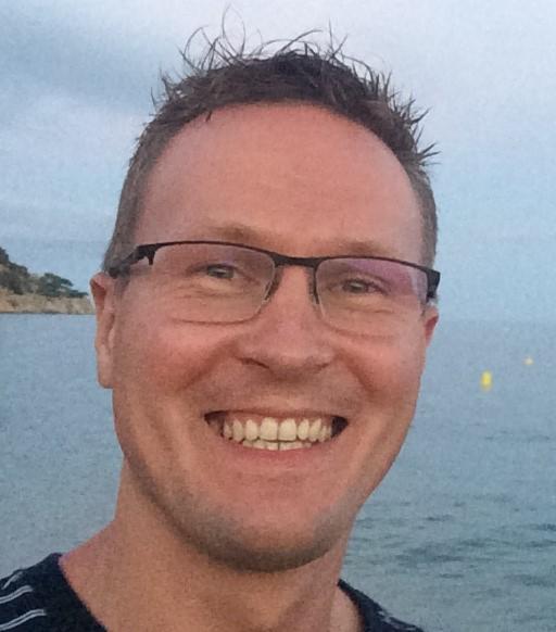 Portret uśmiechniętego mężczyzny w okularach na tle morza i skalistego brzegu w pochmurny dzień. Widoczne białe zęby i krótko ostrzyżone włosy.