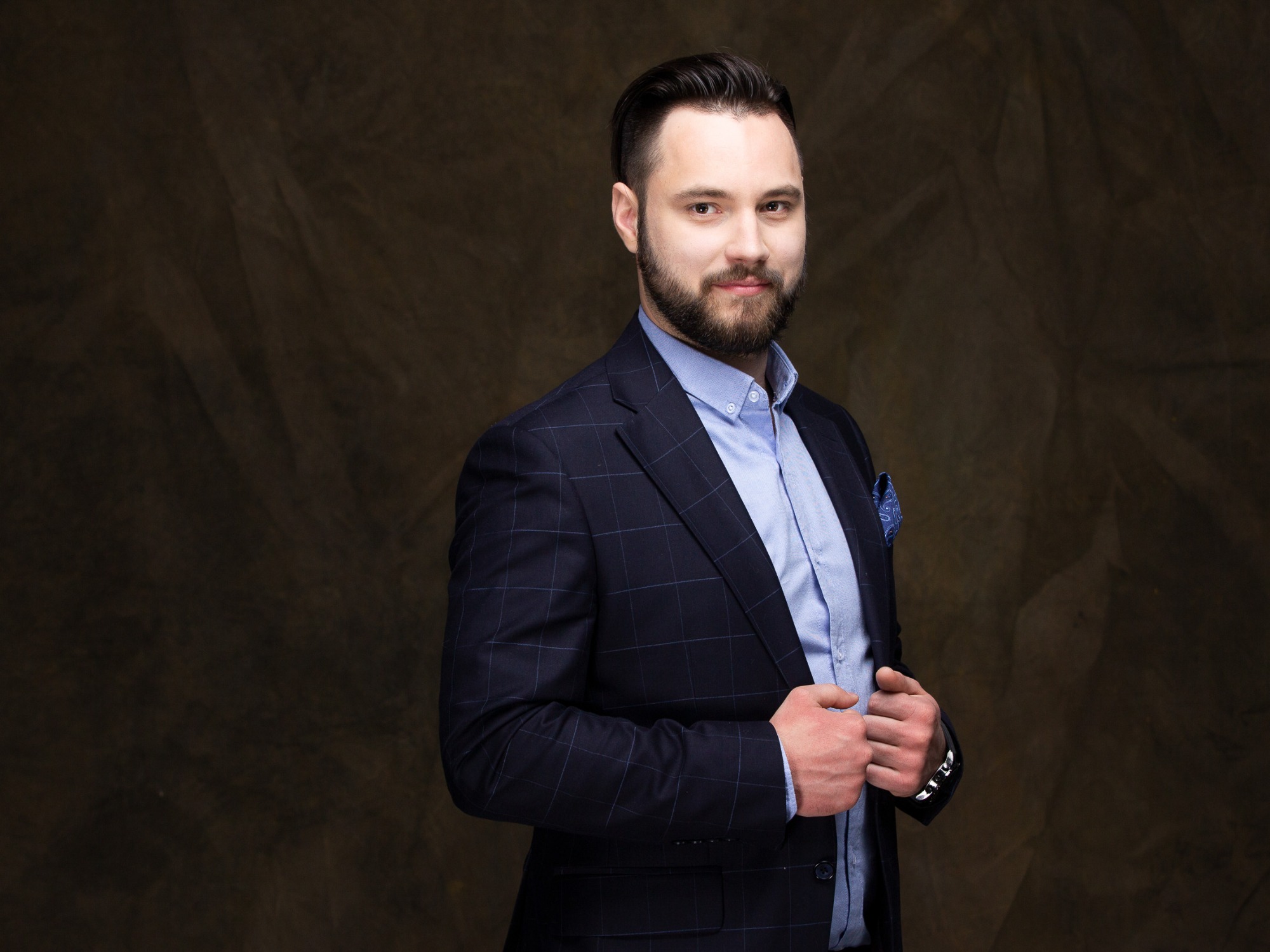 Mężczyzna z brodą w granatowym garniturze w kratę i błękitnej koszuli, pozujący na tle abstrakcyjnego, ciemnego tła.