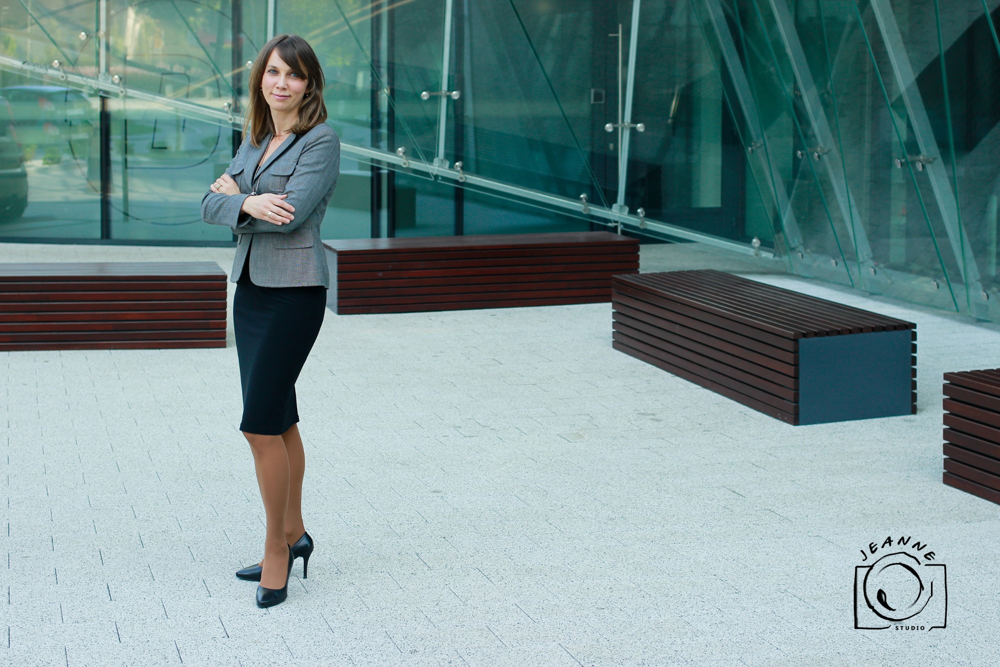 Kobieta w szarym żakiecie i czarnej spódnicy, stojąca na tle nowoczesnego budynku z przeszkloną fasadą i drewnianymi ławkami.