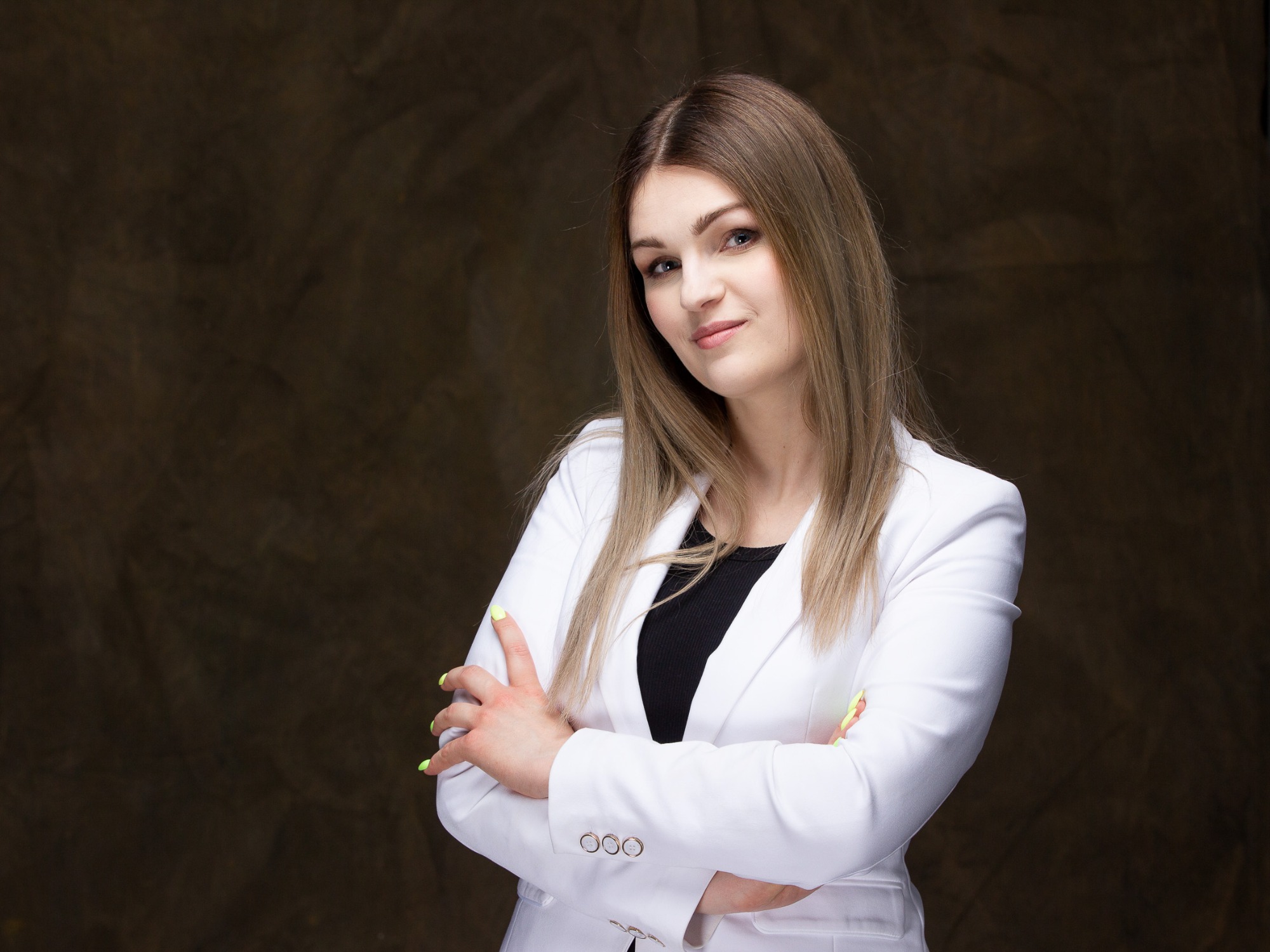Portret kobiety w białym żakiecie i czarnej bluzce, z założonymi rękami i neonowo-zielonym manicure na tle ciemnobrązowego, teksturowanego tła.