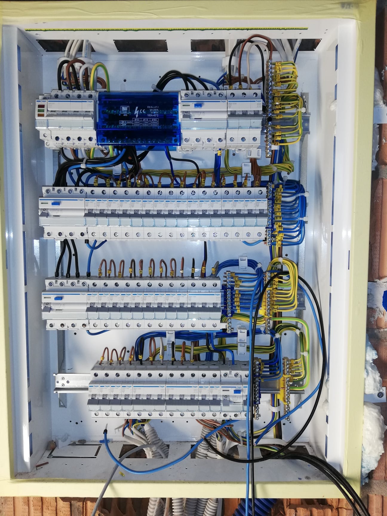 Rozdzielnica elektryczna z zainstalowanymi bezpiecznikami i okablowaniem, widok z góry na białą skrzynkę z żółtą taśmą malarską na krawędziach, z przewodami w różnych kolorach podłączonymi...