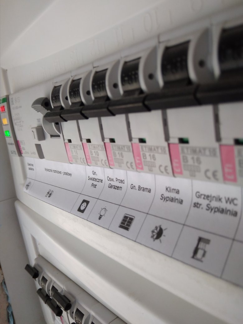 Tablica rozdzielcza z bezpiecznikami i opisami obwodów, w tym gniazda, oświetlenie, garaż, brama, klimatyzacja sypialnia i grzejnik WC, z włącznikami w pozycji włączonej i wyłączonej.