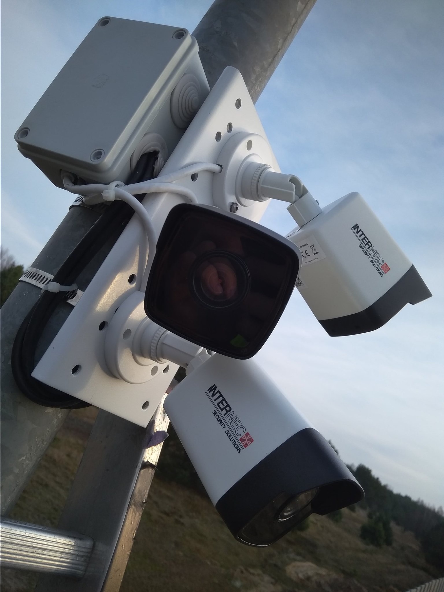 Zestaw kamer monitoringu marki Internec zamontowany na słupie, widoczne okablowanie i puszka instalacyjna, tło stanowi zalesiony teren i pochmurne niebo.