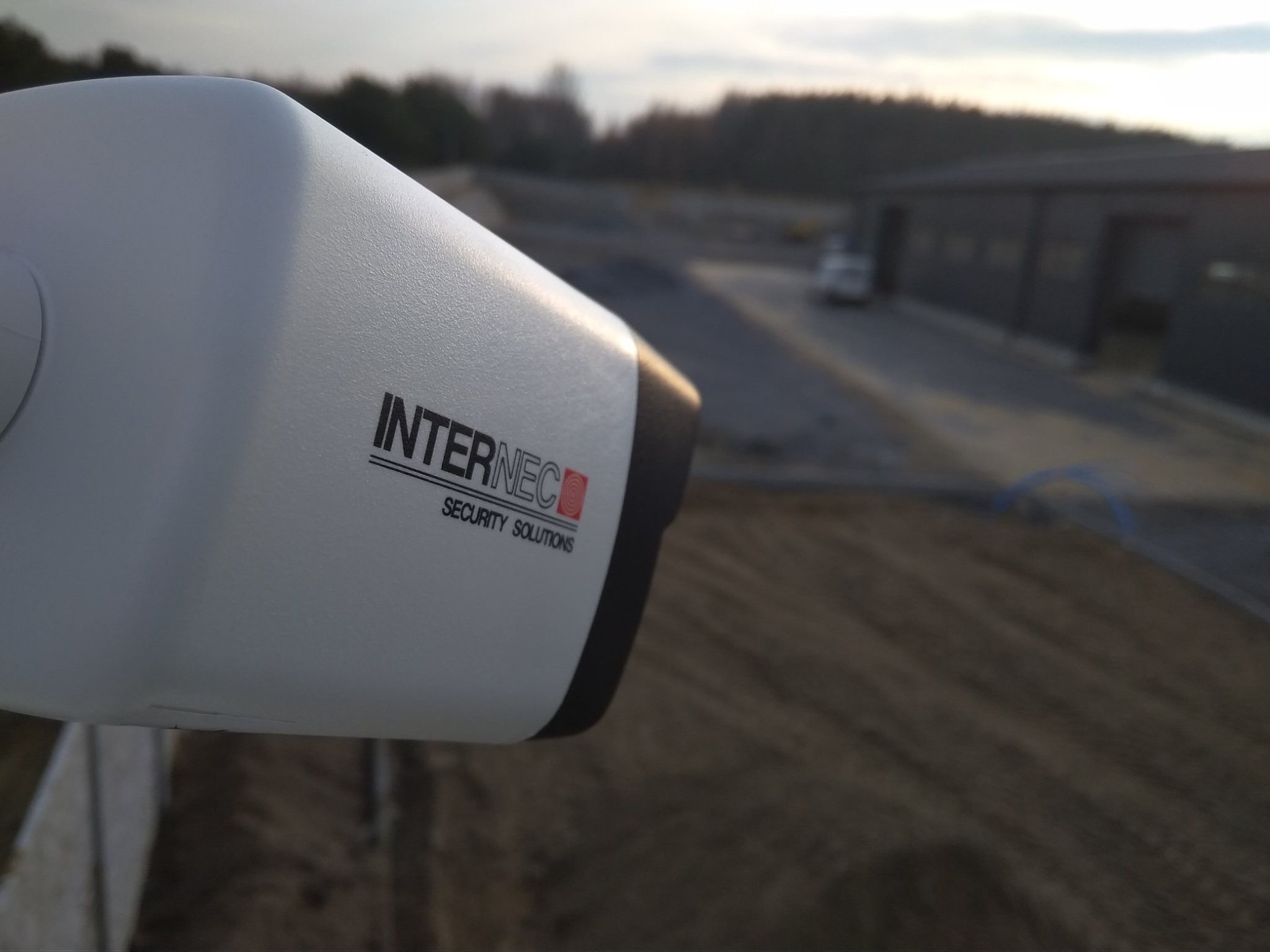 Biała kamera monitoringu z logo Internec Security Solutions, skierowana w stronę parkingu i budynku w tle, sfotografowana o zmierzchu.