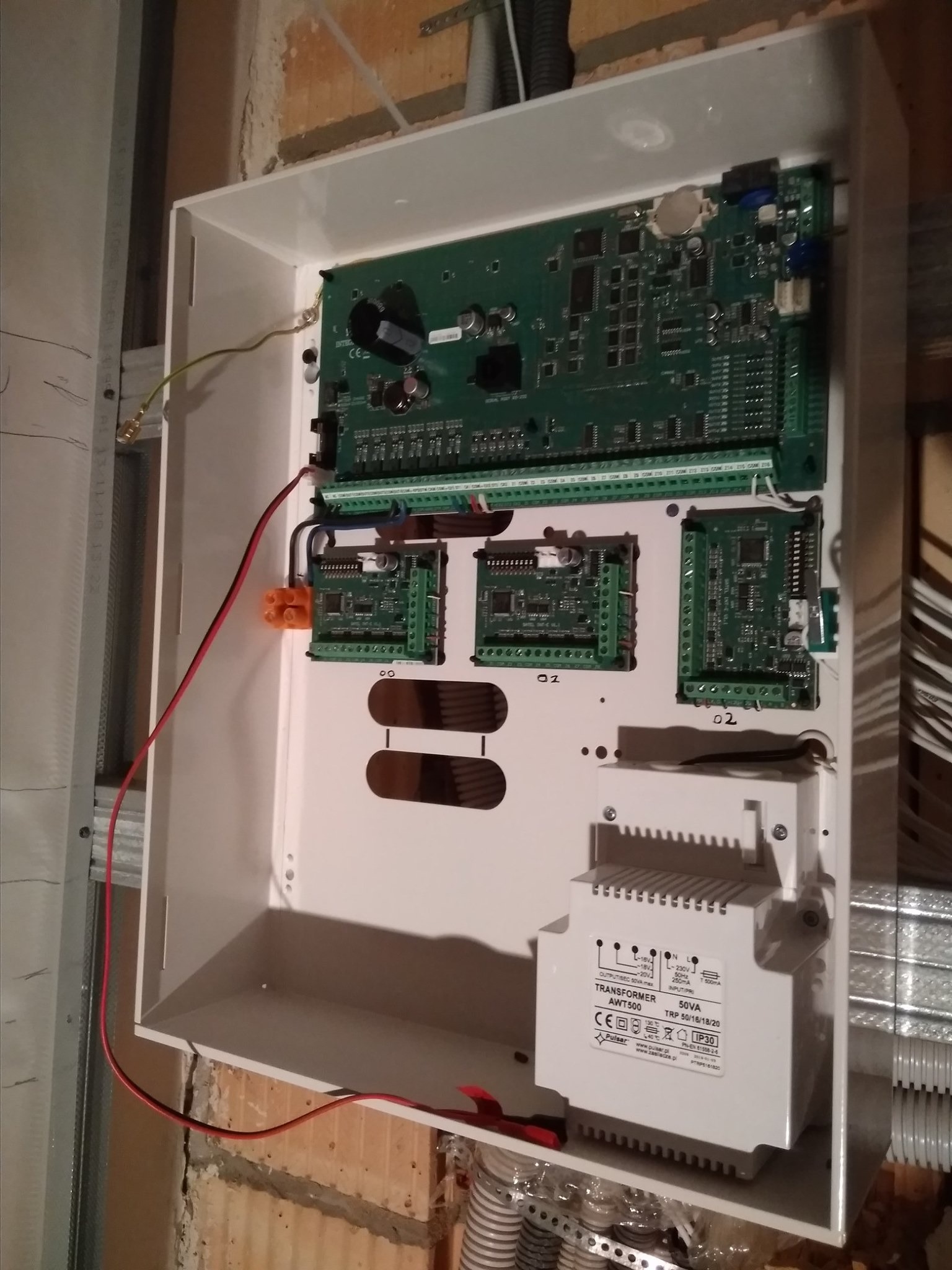 Wewnętrzny widok skrzynki z elektroniką systemu alarmowego podczas montażu, widoczne płytki drukowane, transformator, okablowanie oraz fragment ściany z niedokończonym tynkiem.