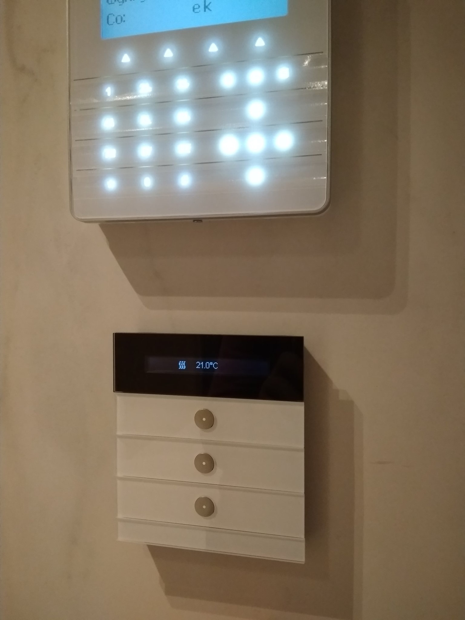 Biały panel alarmowy z podświetlanymi przyciskami oraz regulator temperatury z wyświetlaczem 21 stopni Celsjusza zamontowane na kremowej ścianie.