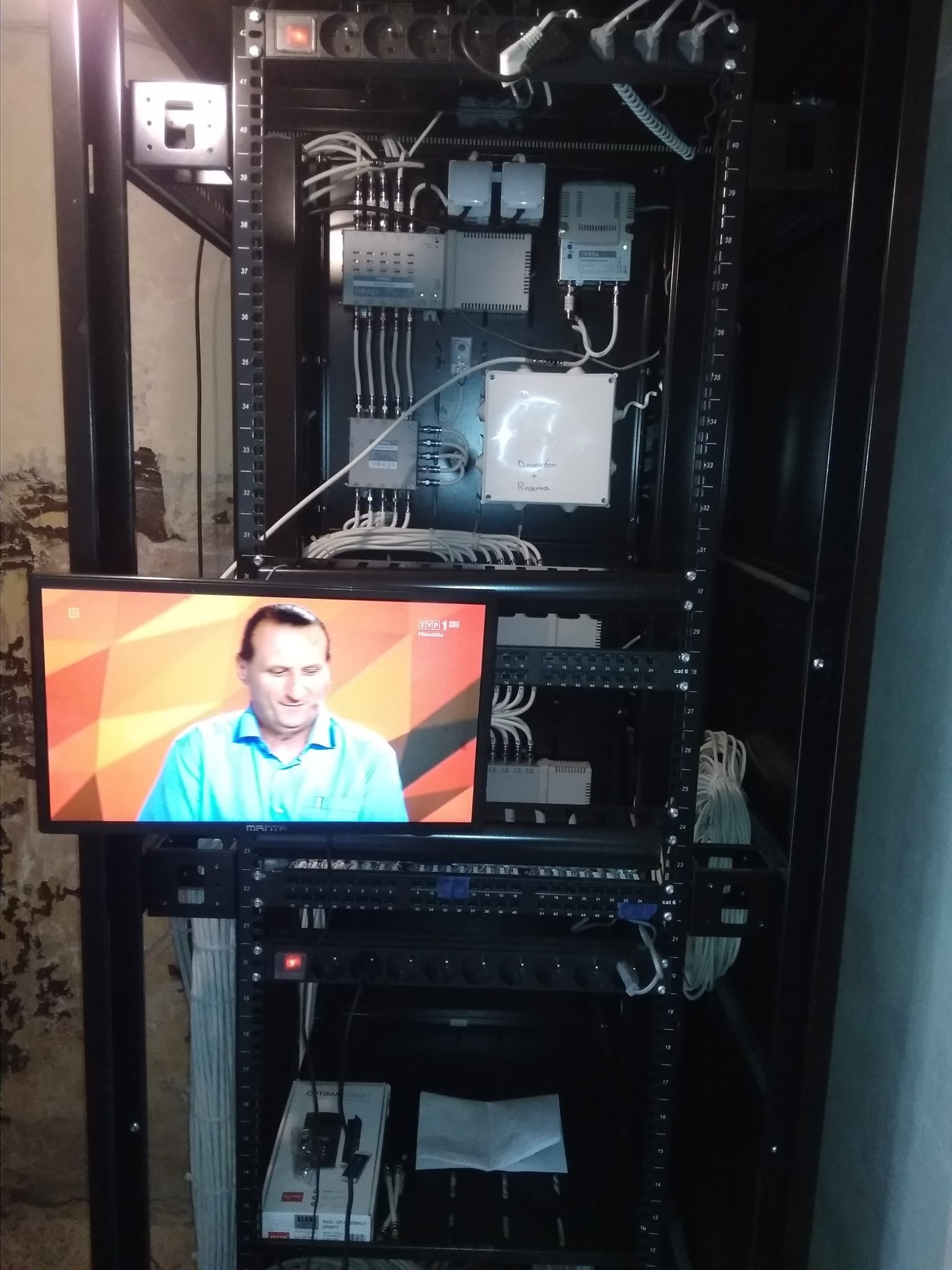 Szafa rack z zainstalowanymi urządzeniami telekomunikacyjnymi, rozdzielnicami i patch panelem, monitor wyświetla program telewizyjny.