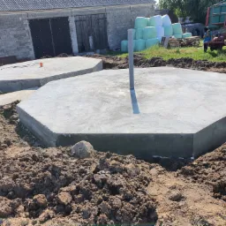 Dwa betonowe, ośmiokątne fundamenty w wykopie, z wystającą rurą pionową na większym fundamencie, w tle budynek gospodarczy i bele sianokiszonki.