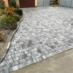 Układanie kostki brukowej z szaro-białych elementów w jodełkę, widoczny fragment nieukończonej nawierzchni z mokrą zaprawą, obrzeża z ciemnej kostki, w tle brama garażowa i rabata z pomarańczowymi...