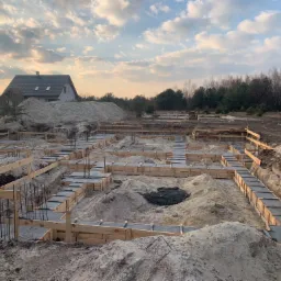 Budowa domu jednorodzinnego: wylane fundamenty z szalunkiem drewnianym i wystającymi prętami zbrojeniowymi, widok na plac budowy z hałdami piasku i domem w tle, pochmurne niebo.