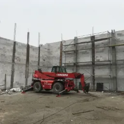 Wznoszenie ścian budynku z betonu komórkowego, widoczny czerwony wózek widłowy Manitou i rusztowania, słupy zbrojeniowe wystają ponad mur.