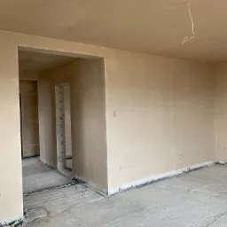 Wnętrze w trakcie prac wykończeniowych: ściany i sufit pokryte świeżym, beżowym tynkiem, widoczne wystające przewody elektryczne, surowa podłoga i otwory drzwiowe z widoczną konstrukcją z pustaków.