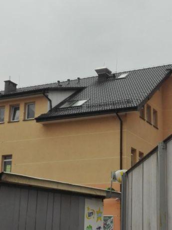 Żółty budynek z ciemnoszarym dachem z dachówki, widoczne okna dachowe i komin, ujęcie z dołu z elementami otoczenia w kadrze.