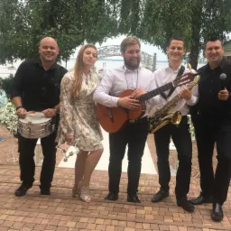 Pięcioosobowy zespół muzyczny z saksofonem, gitarą, bębenkiem i mikrofonem pozuje na tle altanki i dekoracji kwiatowych na zewnątrz.
