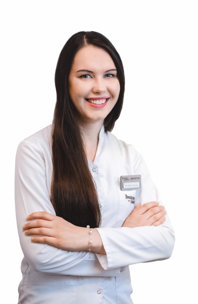 Uśmiechnięta kobieta w białym fartuchu medycznym z plakietką z imieniem i nazwiskiem, skrzyżowanymi rękami i bransoletką z pereł na nadgarstku.