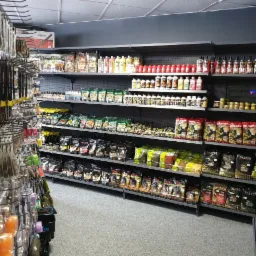 Wnętrze sklepu z artykułami wędkarskimi: półki wypełnione przynętami, zanętami i akcesoriami. Widoczne opakowania marek Traper i Dragon.