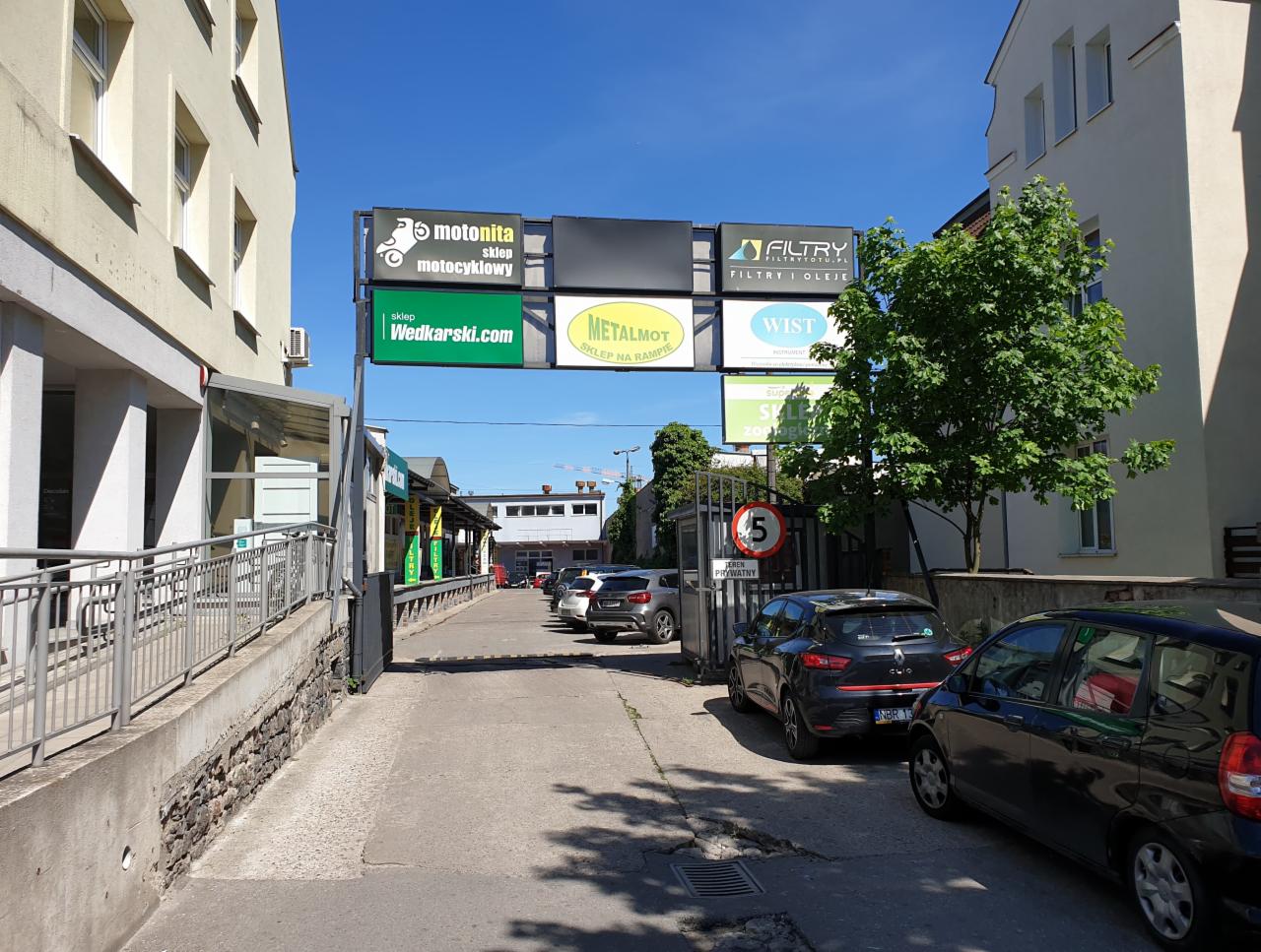 Wjazd na parking z reklamami sklepów motocyklowych, wędkarskich, z filtrami i olejami, oraz sklepów metalowych, widok w kierunku budynków.