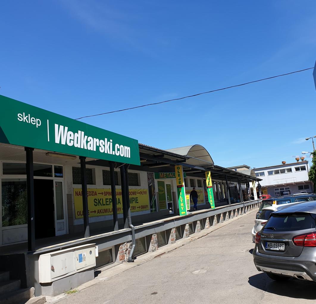 Fasada sklepu Wedkarski.com w Gdańsku, z widocznym szyldem na zielonym tle i zaparkowanymi samochodami wzdłuż ulicy. Na szyldzie widoczny adres strony internetowej. Budynek z licznymi plakatami...