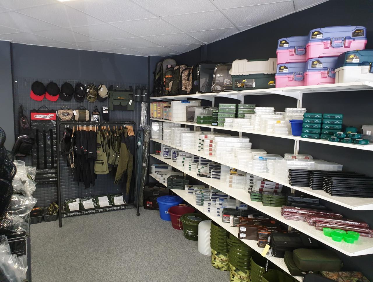 Wnętrze sklepu z artykułami wędkarskimi: półki z pudełkami, torbami i odzieżą wędkarską, w tle ciemna ściana z ekspozycją czapek i akcesoriów.