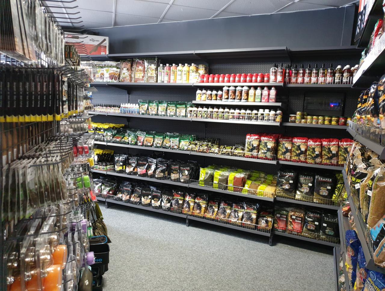 Wnętrze sklepu z artykułami wędkarskimi: półki wypełnione przynętami, zanętami i akcesoriami. Widoczne opakowania marek Traper i Dragon.