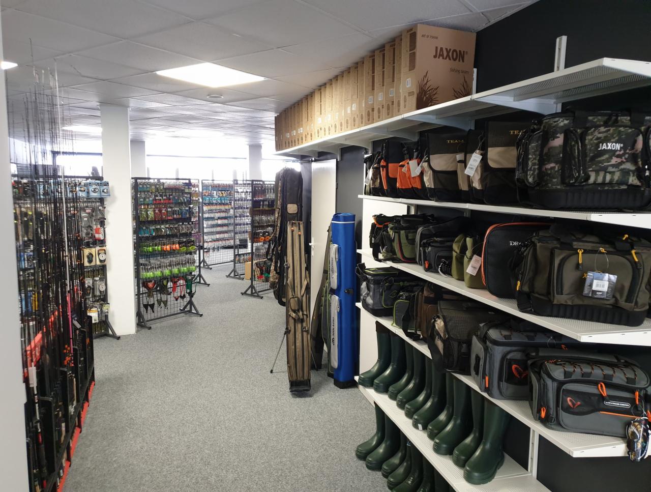 Wnętrze sklepu wędkarskiego z szerokim asortymentem: wędki, przynęty, torby i kalosze na półkach.