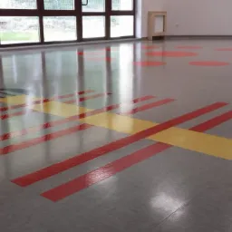 Błyszcząca, szara podłoga z czerwonymi i żółtymi pasami oraz okrągłymi plamami w przedszkolu lub sali zabaw, odbijająca światło z okien.