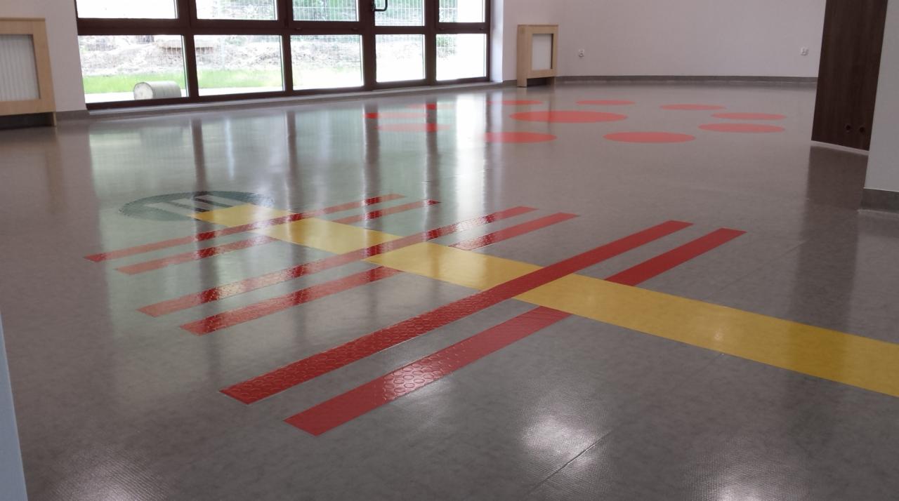 Błyszcząca, szara podłoga z czerwonymi i żółtymi pasami oraz okrągłymi plamami w przedszkolu lub sali zabaw, odbijająca światło z okien.