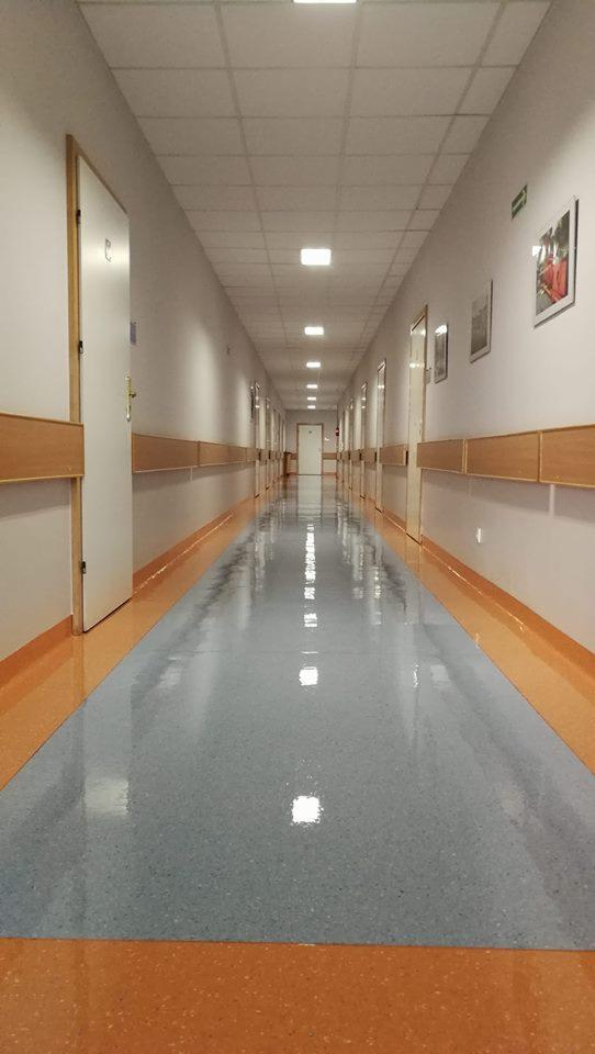 Długi korytarz w budynku użyteczności publicznej z błyszczącą, wyczyszczoną podłogą w odcieniach szarości i pomarańczy, oświetlony rzędami kwadratowych lamp sufitowych, z widocznymi drzwiami...