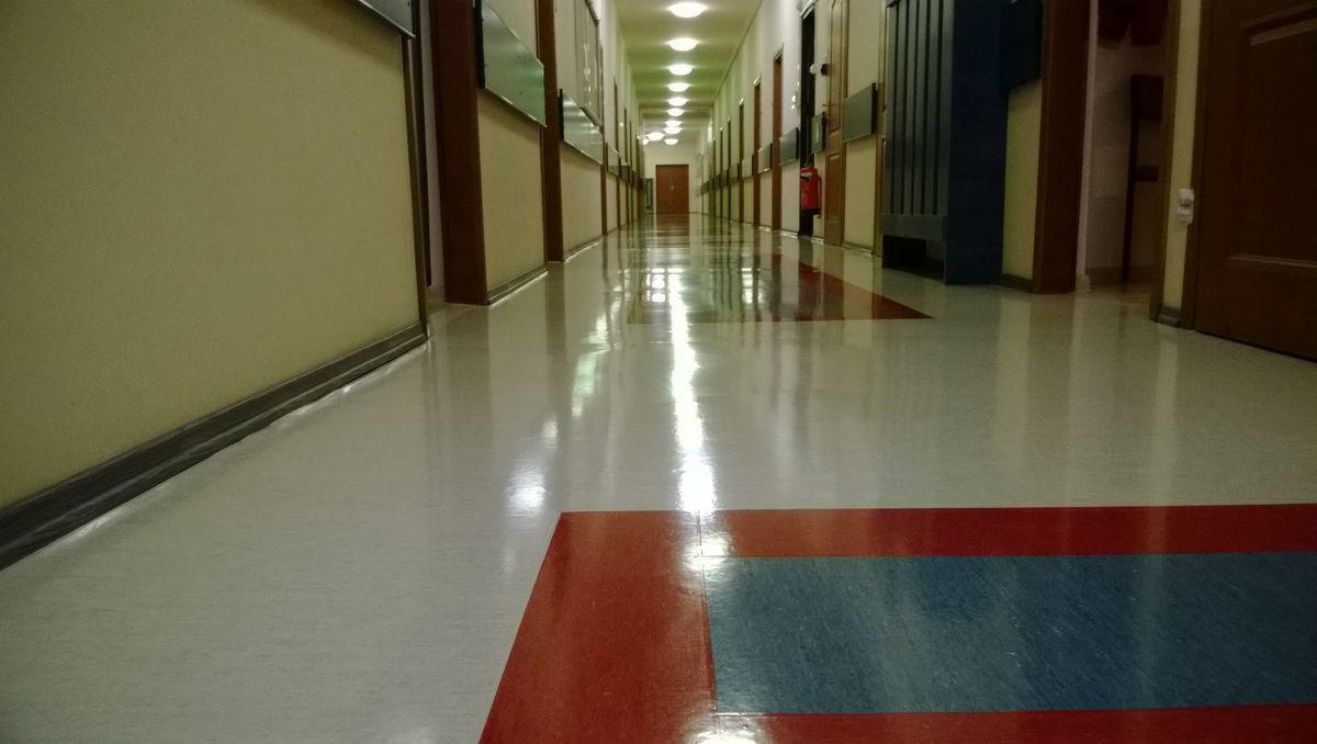 Długi korytarz z wypolerowaną podłogą, widoczne odbicia światła, wzór z czerwonymi i niebieskimi elementami na posadzce, ściany w odcieniach beżu i zieleni, drzwi po obu stronach.