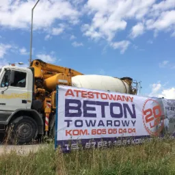 Ciężarówka betoniarka z banerem reklamowym 'Atestowany Beton Towarowy' na tle błękitnego nieba z chmurami.